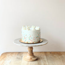 Load image into Gallery viewer, An image of a white buttercream frosting cake decorated with piped rosettes of buttercream on top and colorful sprinkles on the sides. The cake sits atop a marbled cake stand with a wooden base.