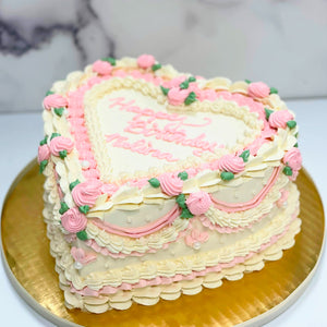 Photo shows a tall heart shaped cake frosted with white buttercream and adorned with intricate buttercream piped swags, rosettes, shell borders and leaves in shades of light pink, ivory and green. Inscription on the center top of the cake reads 'Happy Birthday Malina' in pink piped buttercream.