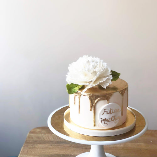 Photo of a white cake with gold ganache dripping down the sides, a white handmade peony on top with green leaves. 