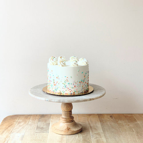 An image of a white buttercream frosting cake decorated with piped rosettes of buttercream on top and colorful sprinkles on the sides. The cake sits atop a marbled cake stand with a wooden base.