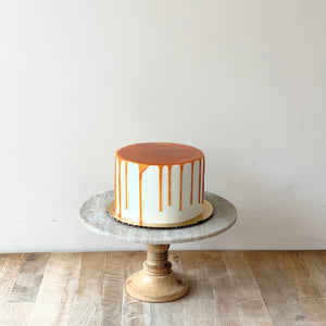 Picture of a white cake with golden brown caramel covering the top of the cake and dripping down the sides. Cake is set atop a wooden and marble cake pedestal.