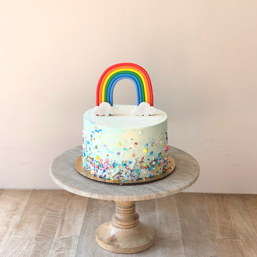 Photo shows a small cake with blue and white watercolor buttercream, multicolored sprinkles and a standing fondant rainbow topper. Cake sits atop a wooden and marble cake pedestal.