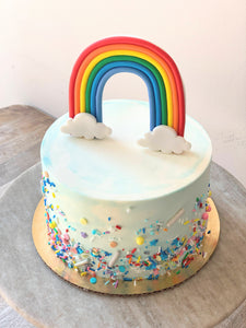 Photo shows a small cake with blue and white watercolor buttercream, multicolored sprinkles and a standing fondant rainbow topper. Cake sits atop a wooden and marble cake pedestal.