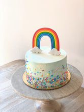 Load image into Gallery viewer, Photo shows a small cake with blue and white watercolor buttercream, multicolored sprinkles and a standing fondant rainbow topper. Cake sits atop a wooden and marble cake pedestal.
