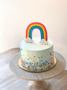 Photo shows a small cake with blue and white watercolor buttercream, multicolored sprinkles and a standing fondant rainbow topper. Cake sits atop a wooden and marble cake pedestal.